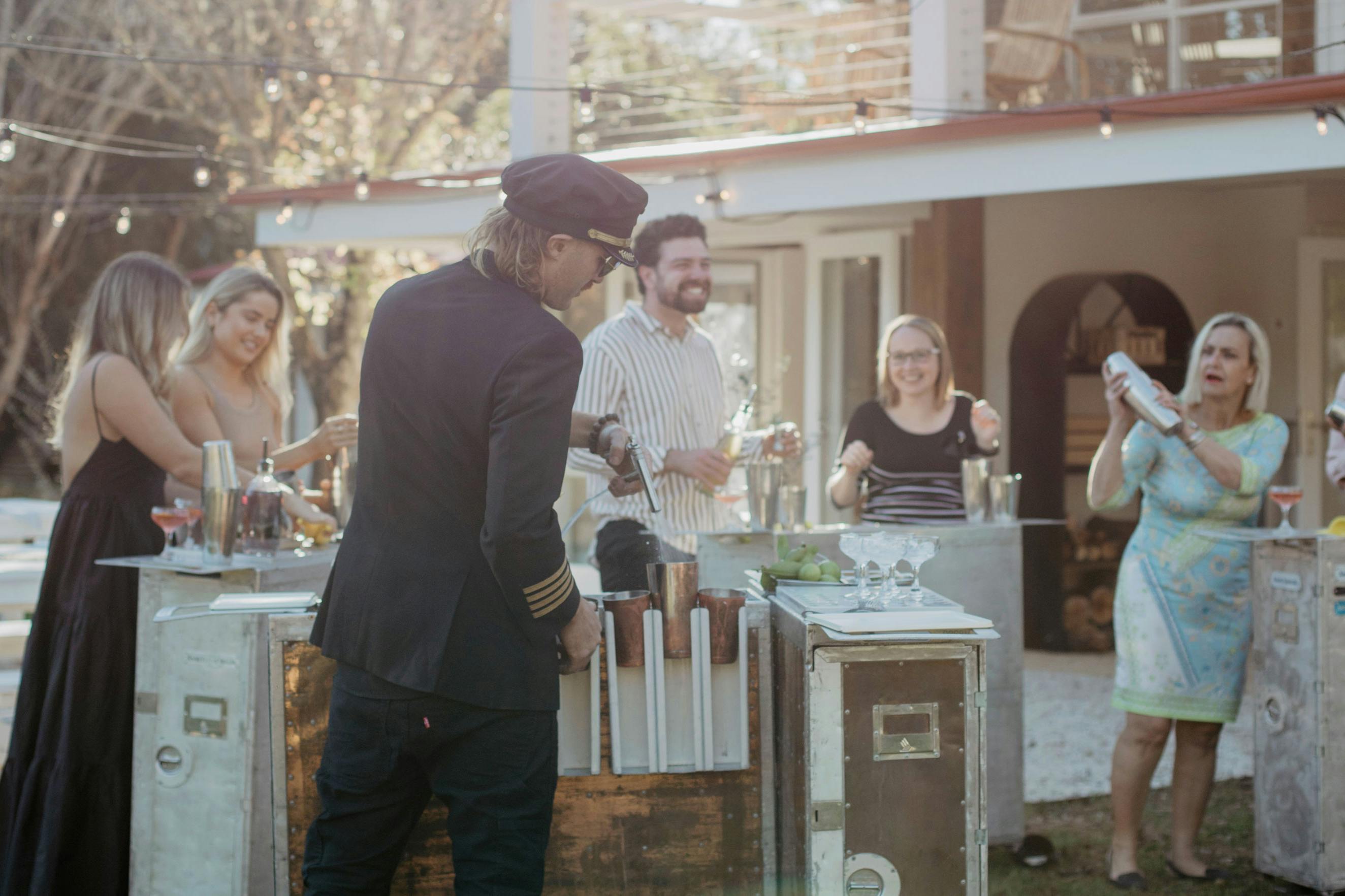 Man with back to camera prepares cocktails for 5 guests at an outdoor garden party